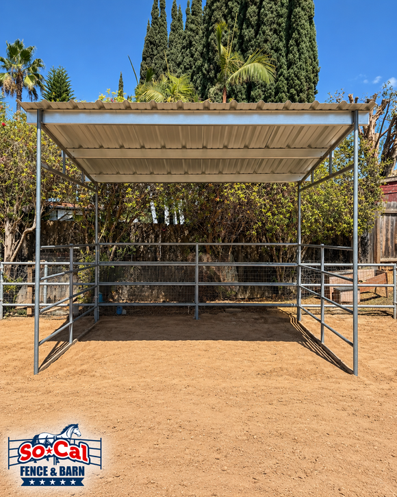 A SoCal Fence and Barn metal horse shelter with a PBR panel roof, set in a sandy outdoor paddock with trees and a blue sky in the background.